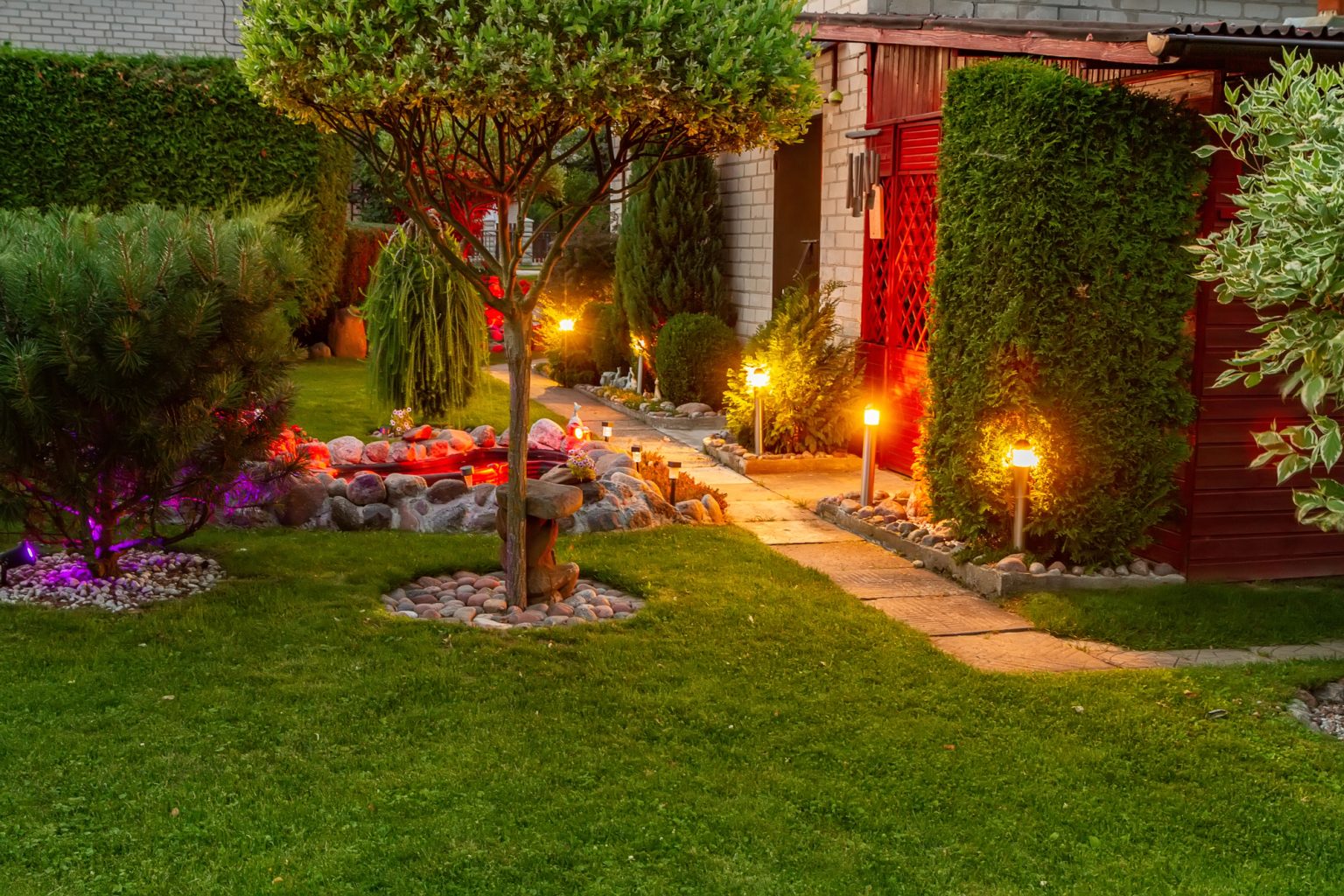 Lawn and garden beds lit by low lights along a stone path at dusk